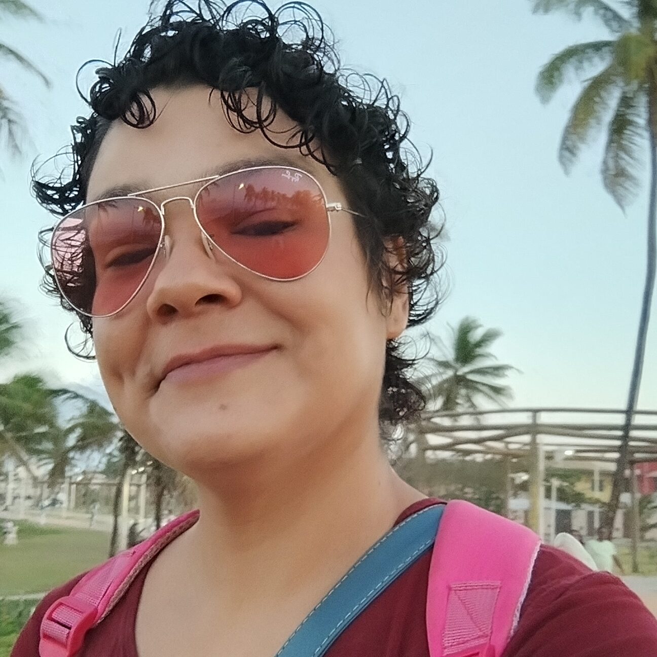 Silvana Machado Valença, mulher de pele clara, cabelos curtos, cacheados e escuros, usando óculos de sol com lentes rosadas e camisa vinho. Ela sorri com expressão acolhedora, carregando uma mochila rosa. A foto foi tirada ao ar livre, em Salvador, com coqueiros e céu claro ao fundo.