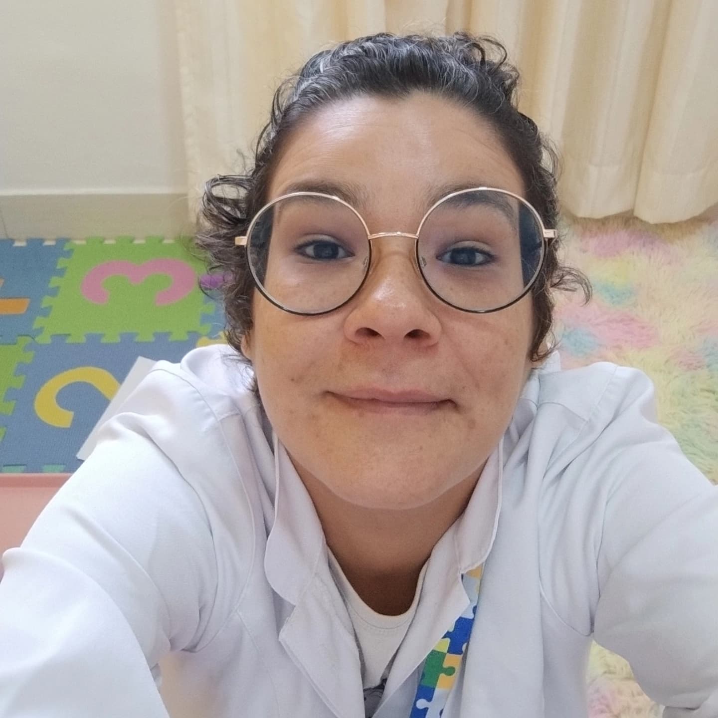 Mulher de pele clara, cabelos castanhos escuros presos e cacheados, usando óculos de armação redonda metálica. Ela veste um jaleco branco e sorri levemente enquanto tira uma selfie de cima para baixo. No pescoço, usa um cordão colorido com estampa de peças de quebra-cabeça, símbolo associado ao Transtorno do Espectro Autista (TEA). Ao fundo, o ambiente tem um tapete emborrachado com números coloridos, um tapete felpudo em tons pastel e cortinas claras, indicando um espaço acolhedor, lúdico e voltado para o atendimento infantil.
