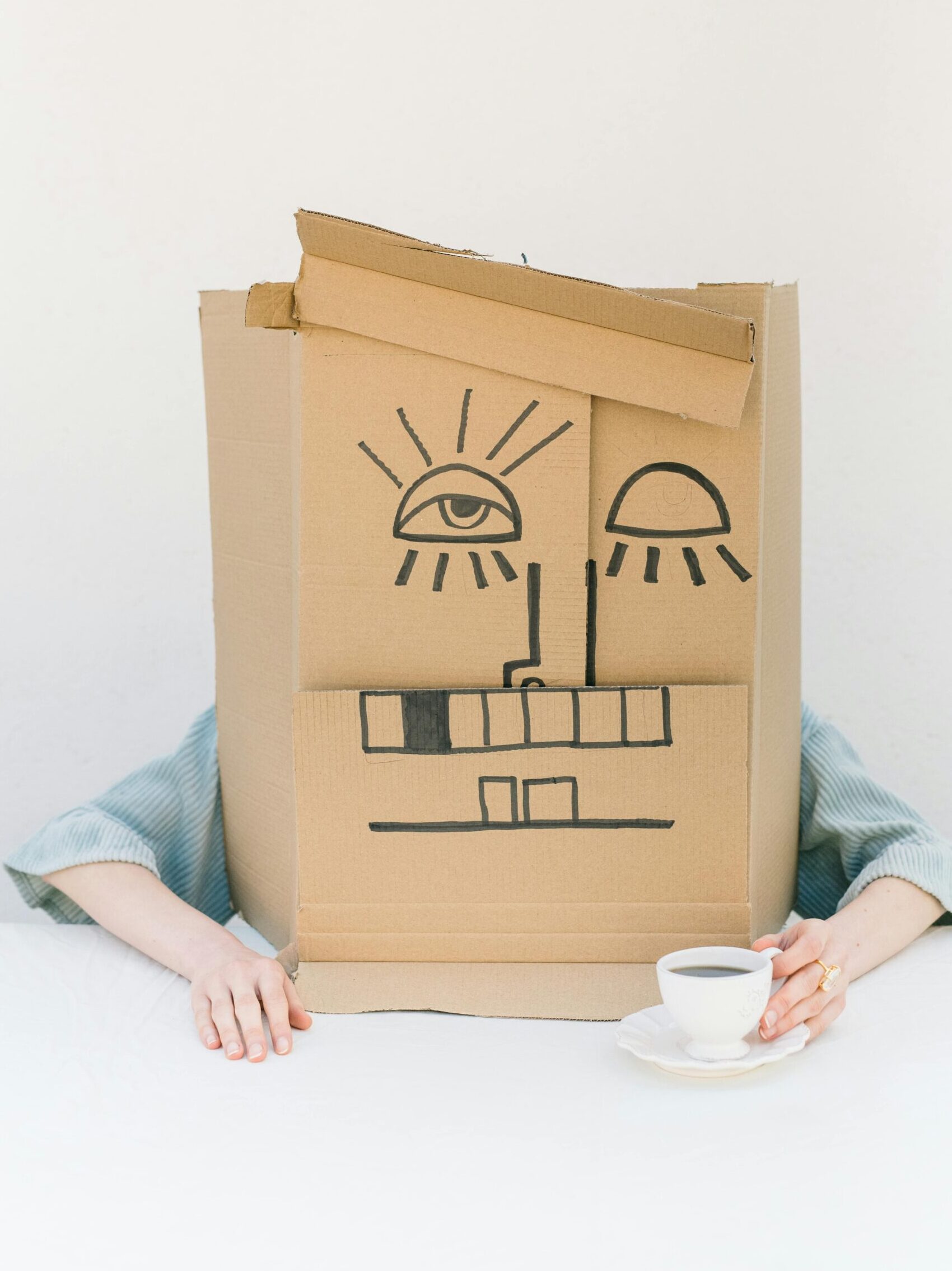 Person wearing a cardboard box mask, sipping coffee at a minimalist setup.