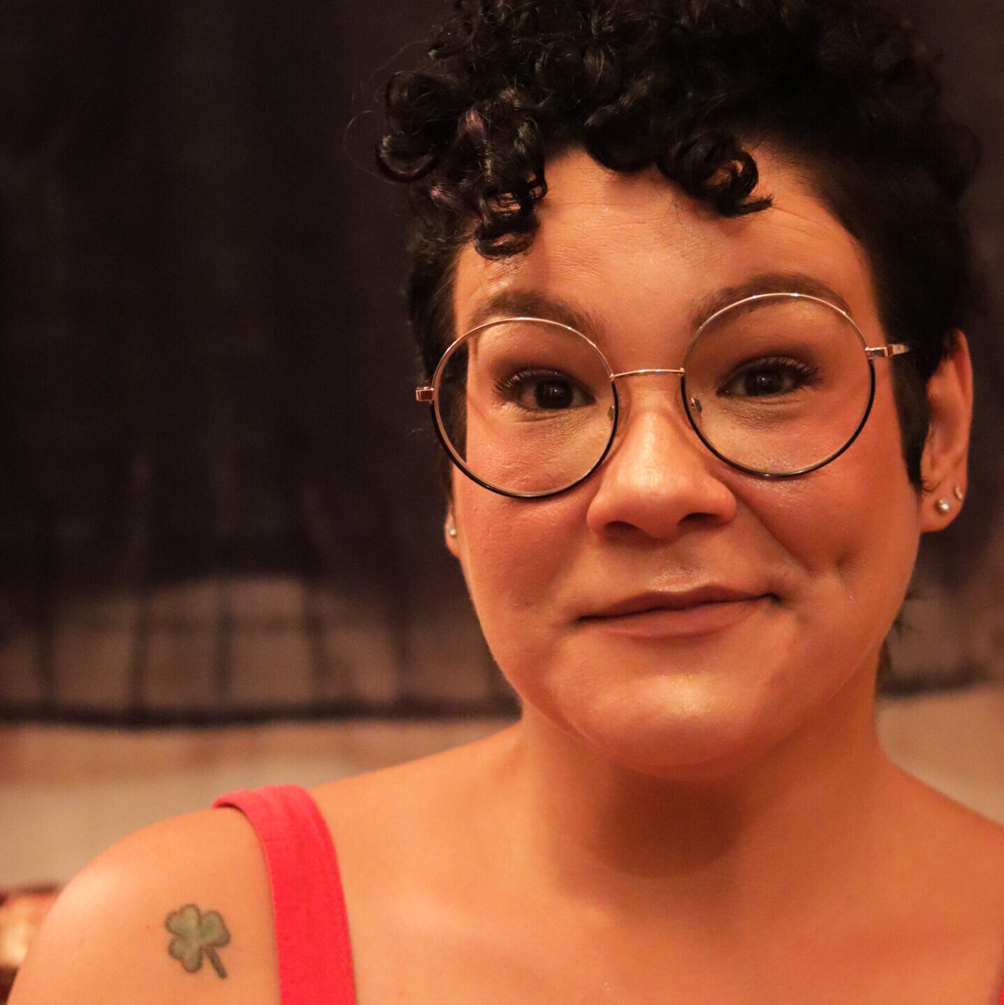 Mulher de cabelos cacheados curtos e escuros, usando óculos redondos e vestido vermelho, com expressão serena. Ela tem covinhas marcantes e uma tatuagem de trevo de três folhas no ombro esquerdo. O fundo é escuro e desfocado, com iluminação quente e suave. Psicóloga feminista, autista e gestalt-terapeuta.