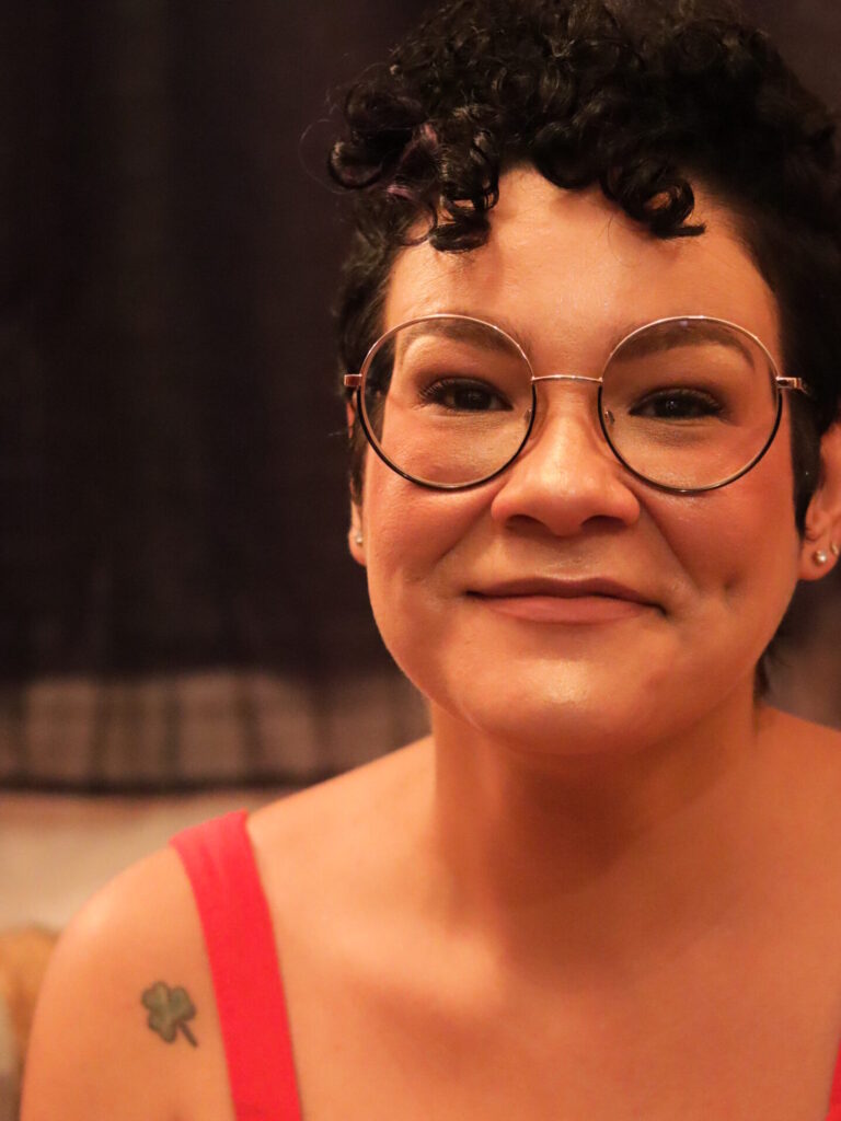 Mulher sorridente com cabelos cacheados curtos e escuros, usando óculos redondos e vestido vermelho. Ela tem uma tatuagem de trevo de três folhas no ombro esquerdo. A iluminação é quente e o fundo desfocado, criando um clima acolhedor e íntimo.