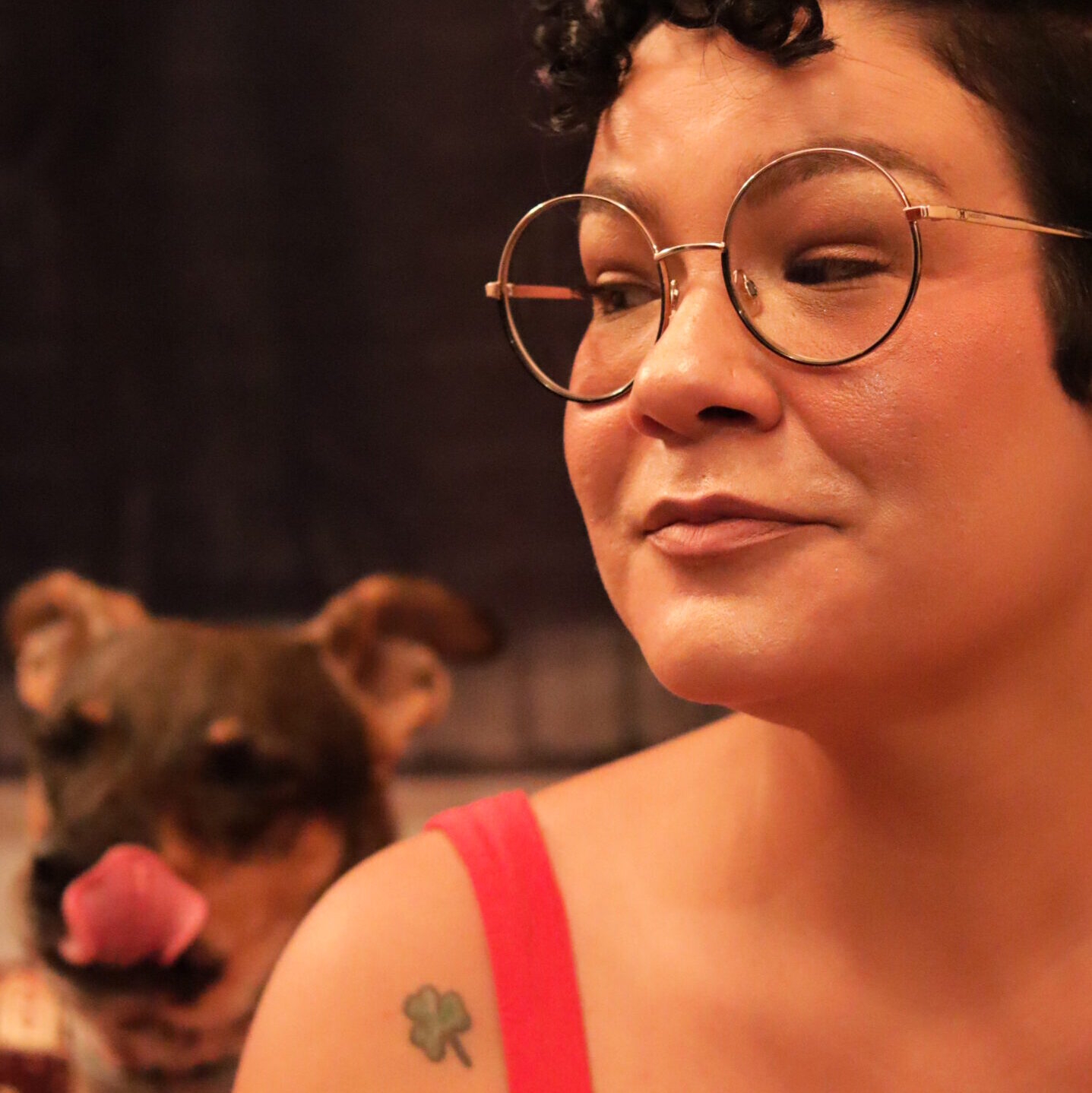 Mulher de cabelos curtos e cacheados, usando óculos redondos e vestido vermelho, olha de lado com expressão divertida. Ao fundo, um cachorro aparece lambendo o focinho, adicionando um toque de humor e espontaneidade à cena. A tatuagem de trevo de três folhas no ombro esquerdo é visível.