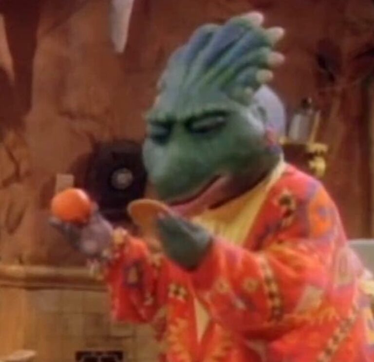 Personagem dinossauro verde, com crista na cabeça, usando um casaco colorido em tons de vermelho, laranja e amarelo, observa atentamente uma bolacha em uma mão e uma fruta laranja (possivelmente uma tangerina) na outra, dentro de uma cozinha com paredes em tom terroso ao fundo.